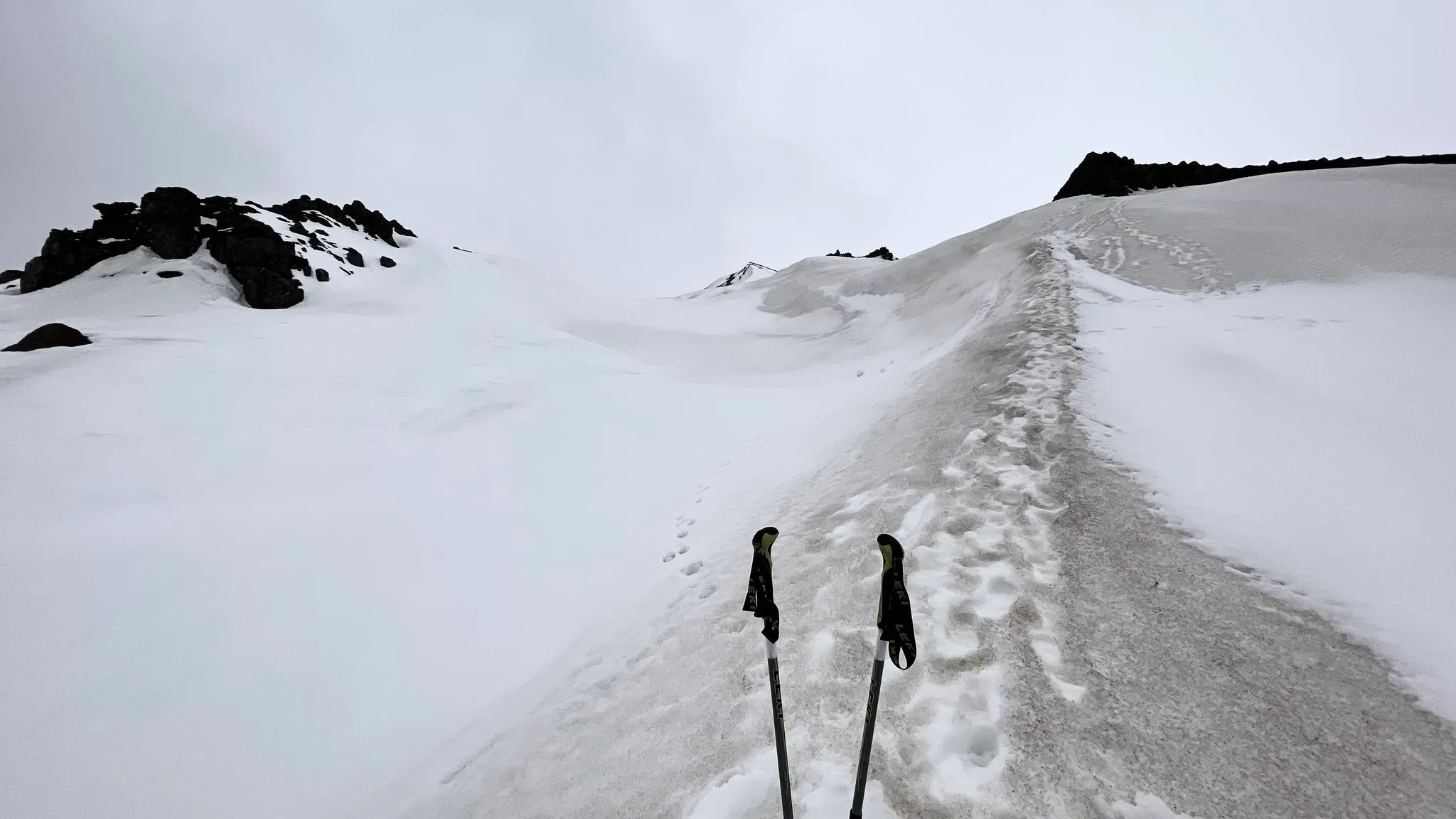 A boot-track winding across a summer snowfield, walking poles planted beside it